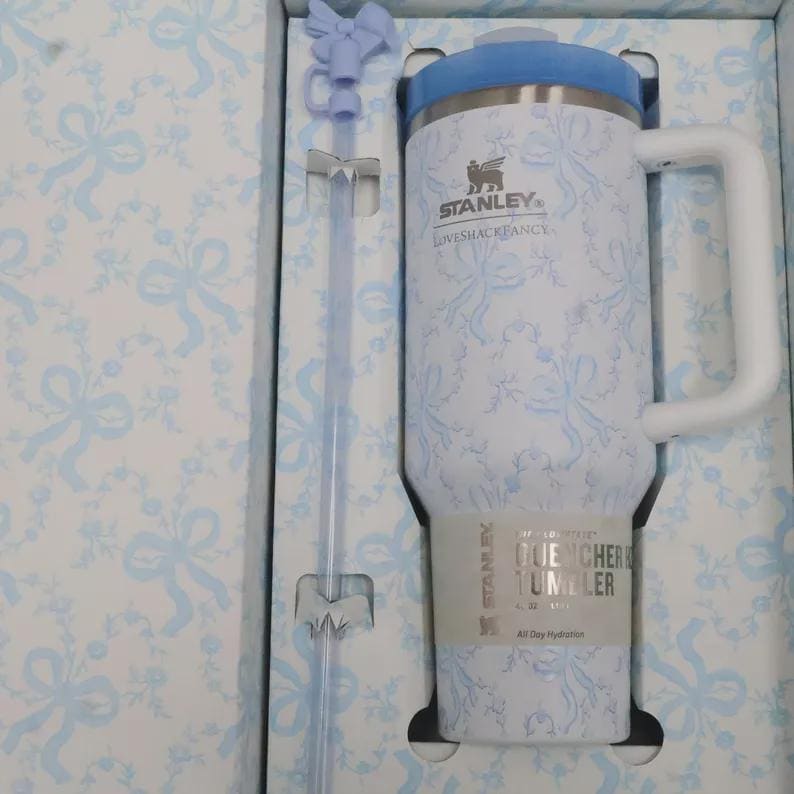 May include: A white and blue Stanley Quencher tumbler with a handle and straw, presented in a matching patterned box. The tumbler features a light blue lid and a floral bow design. The box and straw also have the same bow pattern.