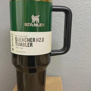 K&ouml;nnte beinhalten: Ein gr&uuml;ner und schwarzer Stanley Quencher H2.0 Tumbler mit schwarzem Griff und Deckel. Der Becher hat das Stanley-Logo und den Text "QUENCHER H2.0 TUMBLER" auf einem wei&szlig;en Etikett. Er fasst 1,18 L.