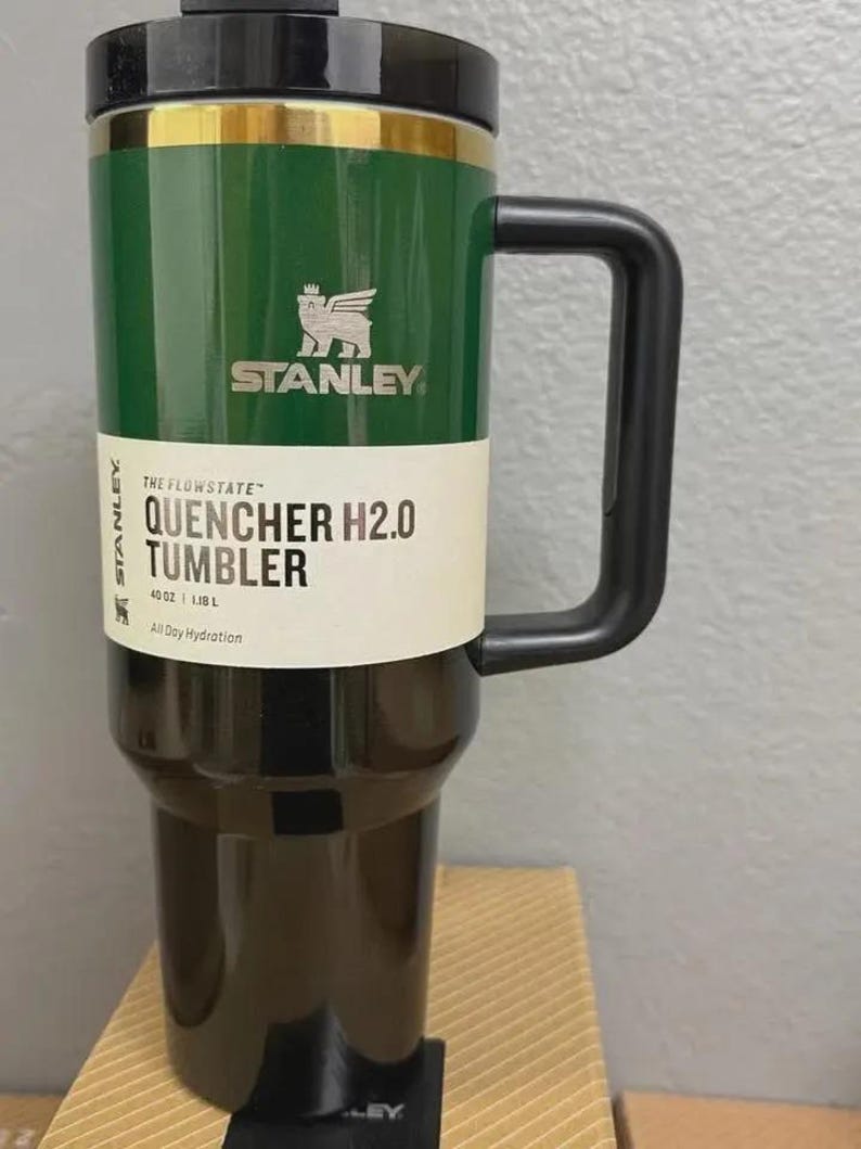 K&ouml;nnte beinhalten: Ein gr&uuml;ner und schwarzer Stanley Quencher H2.0 Tumbler mit schwarzem Griff und Deckel. Der Becher hat das Stanley-Logo und den Text "QUENCHER H2.0 TUMBLER" auf einem wei&szlig;en Etikett. Er fasst 1,18 L.