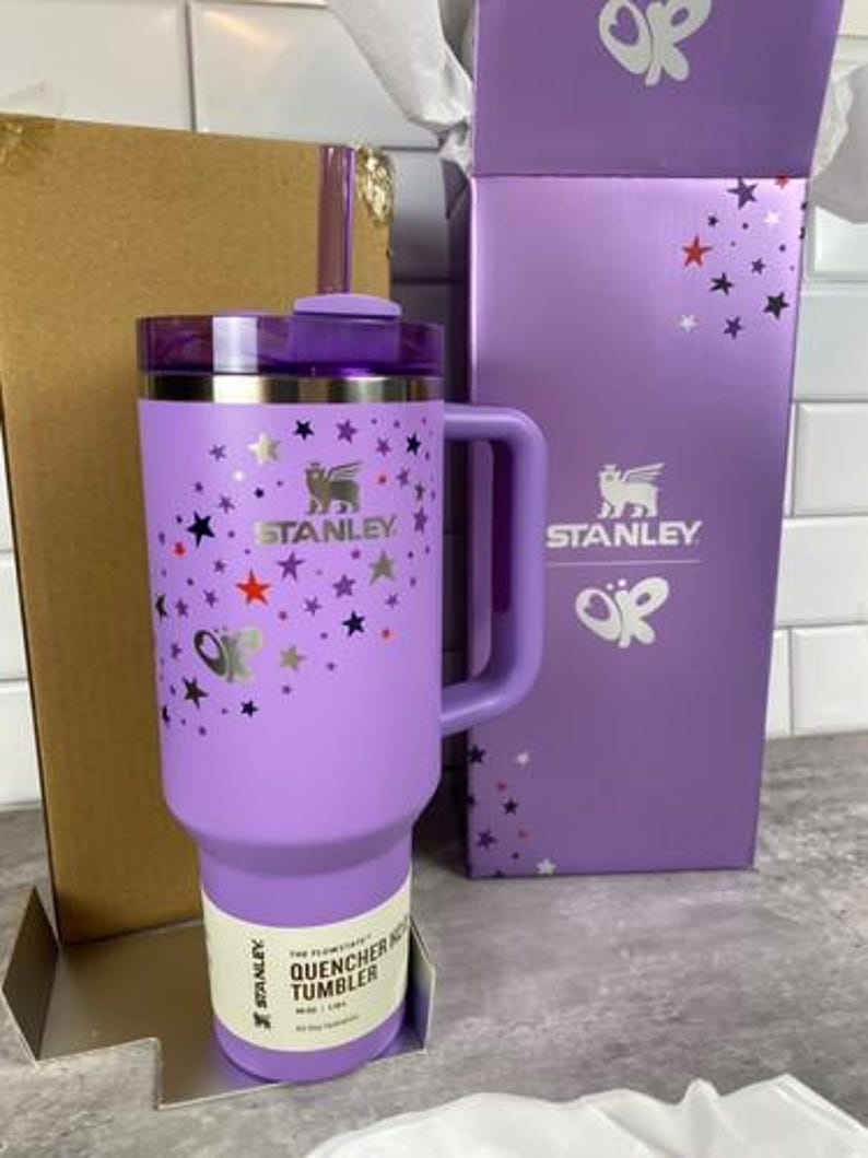 Może przedstawiać: Lawendowy kubek Stanley Quencher H2.0 FlowState z uchwytem, pokrywką i słomką. Kubek jest ozdobiony srebrnymi, czerwonymi i czarnymi gwiazdami. Pudełko jest lawendowe z logo Stanley i wzorami gwiazd.