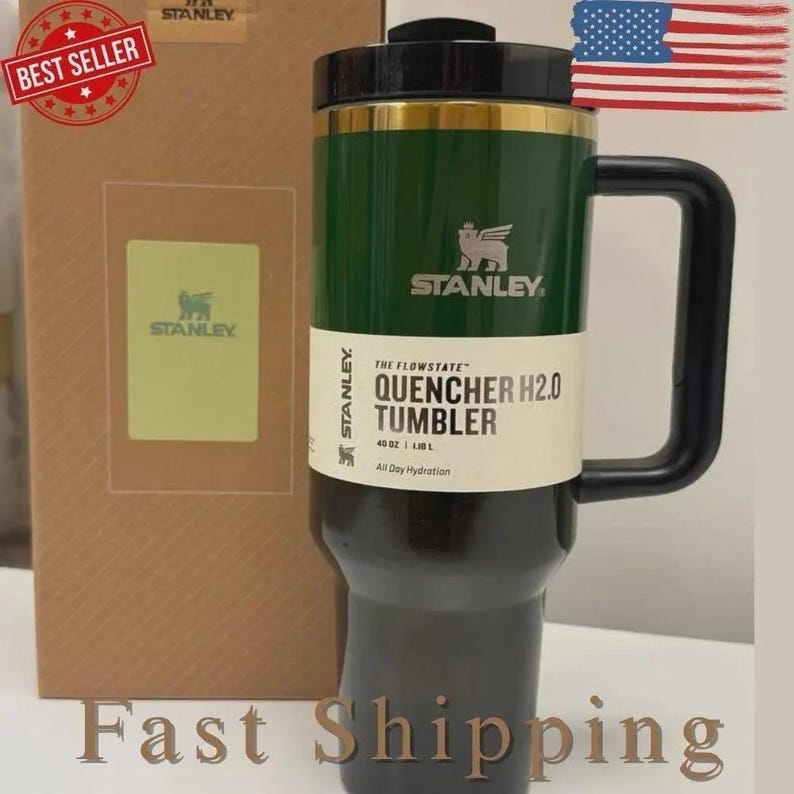 K&ouml;nnte beinhalten: Gr&uuml;ner und schwarzer Stanley Quencher H2.0 Becher mit schwarzem Griff. Der Becher hat ein goldenes Band und das Stanley-Logo. Der Text auf dem Becher lautet "QUENCHER H2.0 TUMBLER" und "40 OZ | 1,18 L".