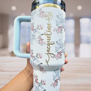 Pu&ograve; includere: Bicchiere isolante bianco con manico e coperchio azzurro. Il bicchiere &egrave; decorato con un motivo floreale di rose rosa e fiocchi blu. Il nome "Jacqueline" &egrave; scritto in oro sul lato.