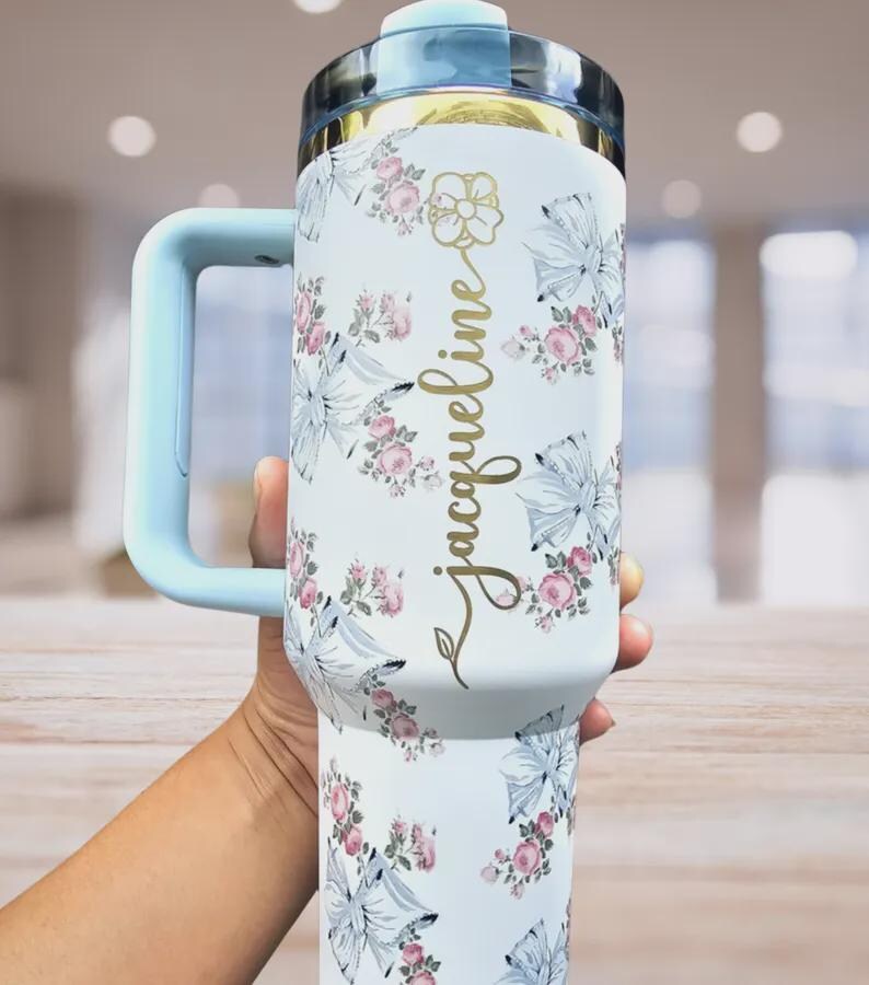 Pu&ograve; includere: Bicchiere isolante bianco con manico e coperchio azzurro. Il bicchiere &egrave; decorato con un motivo floreale di rose rosa e fiocchi blu. Il nome "Jacqueline" &egrave; scritto in oro sul lato.