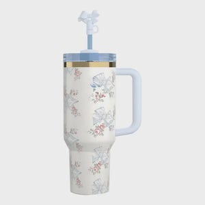Pu&ograve; includere: Un bicchiere termico bianco con un motivo floreale e a fiocco. Il bicchiere ha un coperchio, un manico e una cannuccia azzurri con un ornamento a forma di fiocco. Una fascia dorata adorna la parte superiore del bicchiere.