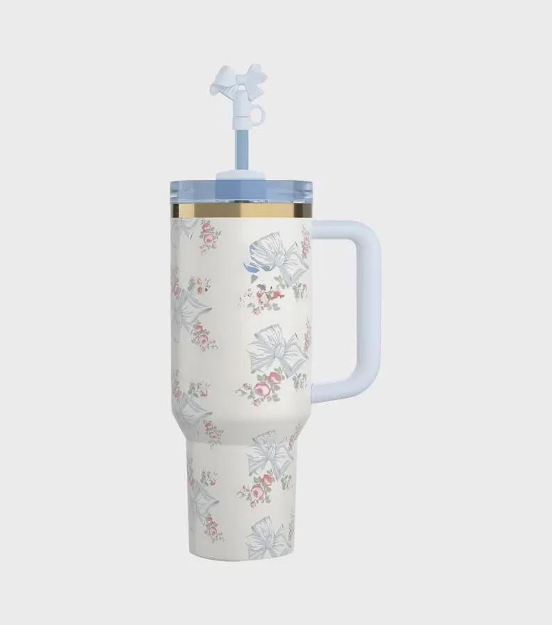 Pu&ograve; includere: Un bicchiere termico bianco con un motivo floreale e a fiocco. Il bicchiere ha un coperchio, un manico e una cannuccia azzurri con un ornamento a forma di fiocco. Una fascia dorata adorna la parte superiore del bicchiere.