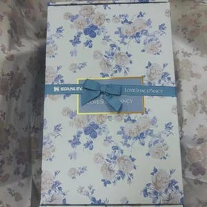 May include: A rectangular gift box featuring a floral design in white, blue, and beige. A blue ribbon with a bow is tied around the box, with the text "STANLEY" and "LOVESHACKFANCY" displayed.