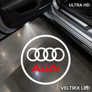 May include: A white illuminated Audi logo projected onto a gray surface from a car door light. The word "Audi" is in red. The text "ULTRA HD" and "VELTRIX LED" are also visible.