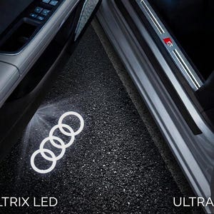May include: An open car door reveals a white Audi logo projected onto the asphalt. The door sill features an "S" emblem. The text "VELTRIX LED" and "ULTRA HD" are visible.