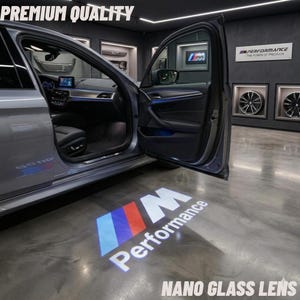 May include: A gray car with the door open, displaying a projected logo on the floor. The logo is the BMW M Performance emblem in white, red, and blue. The text "PREMIUM QUALITY" and "NANO GLASS LENS" are also visible.