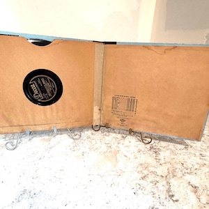 May include: An open vintage Decca record album folder with a light blue exterior and tan interior sleeves. The left side holds a black vinyl record with a Decca label, while the right side features a printed list of other Decca record releases.