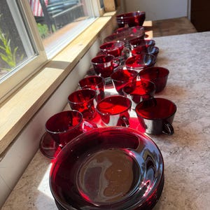 以下が含まれることがあります： ルビーレッドのガラス製食器コレクション。持ち手付きのティーカップと、それに対応するソーサーが複数含まれています。半透明の赤いガラス製品は列状に並べられており、滑らかで光沢のある仕上げと、深く鮮やかな深紅の色合いが特徴です。