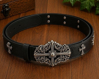 May include: A black belt featuring a large, ornate silver-toned metal buckle with intricate scrollwork and a central cross design. The belt strap is adorned with smaller silver-toned cross-shaped metal studs and circular rivets along the band.