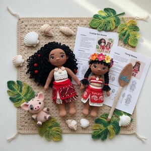 May include: Handmade crochet dolls of Moana and a pig, with a leaf and seashell arrangement. The dolls have black hair, red skirts, and white tops. The pig is pink and gray. Crochet leaves and seashells are scattered around the dolls.