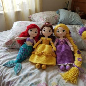 May include: Three handmade crochet dolls: Ariel with red hair and a teal tail, Belle in a yellow dress, and Rapunzel with long blonde hair and a purple dress. The dolls are on a floral patterned bedspread.