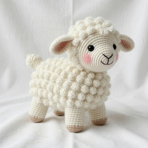May include: A handmade, off-white crochet sheep with a textured body. The sheep has a smiling face with pink cheeks, black eyes, and tan hooves. The ears are pink inside. The sheep is standing on a white surface.