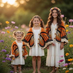 Puede incluir: Tres niñas con vestidos blancos y cárdigans de ganchillo con forma de alas de mariposa naranja y negra, de pie en un campo de flores coloridas. Los cárdigans tienen cierres de botón y ribetes color crema. Una niña lleva una corona de flores.