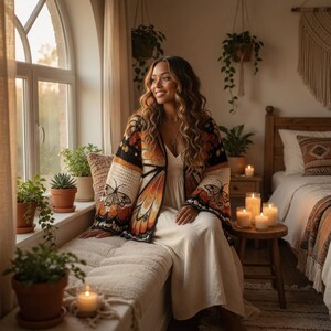 May include: A woman wearing a cream-colored dress and a butterfly-patterned cardigan sits by a window. The cardigan features black, orange, and cream colors. The room is decorated with plants, candles, and a wooden bed frame.