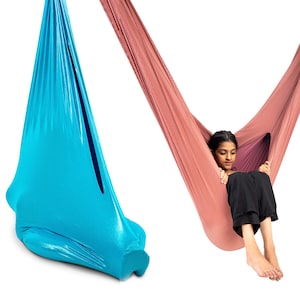 May include: A bright blue sensory swing made of stretchy fabric hanging vertically, alongside a dusty rose sensory swing made of textured fabric with a person sitting inside, wearing black trousers, suspended against a plain white background.