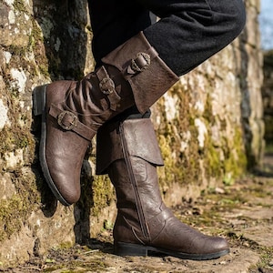May include: A pair of brown mid-calf boots featuring a textured finish, a folded cuff at the top, and two decorative straps with antique brass-toned circular buckles. The boots include a side zipper closure and a low heel for a functional design.