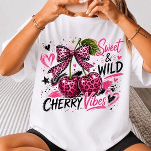 Może przedstawiać: Biały t-shirt z grafiką przedstawiającą dwie wiśnie w różowy wzór panterki. Wiśnie są połączone pasującą kokardą w różowy wzór panterki. Napis głosi Sweet & WILD CHERRY Vibes, a całość uzupełniają różowe serca, czarne gwiazdki i pociągnięcia pędzla.