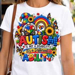 May include: A white t-shirt featuring a colorful graphic of a gnome holding a heart-shaped puzzle piece, sunflowers, butterflies, and a rainbow. The text reads: AUTISM IS NOT A DISABILITY It's a Different ABILITY, decorated with puzzle patterns.