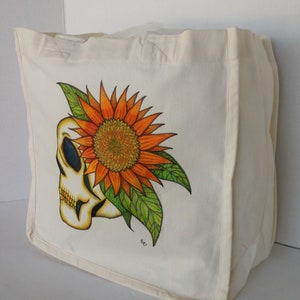 May include: A white canvas tote bag with a hand-painted design of a skull with a sunflower growing out of the top of its head. The sunflower is orange and yellow with green leaves.
