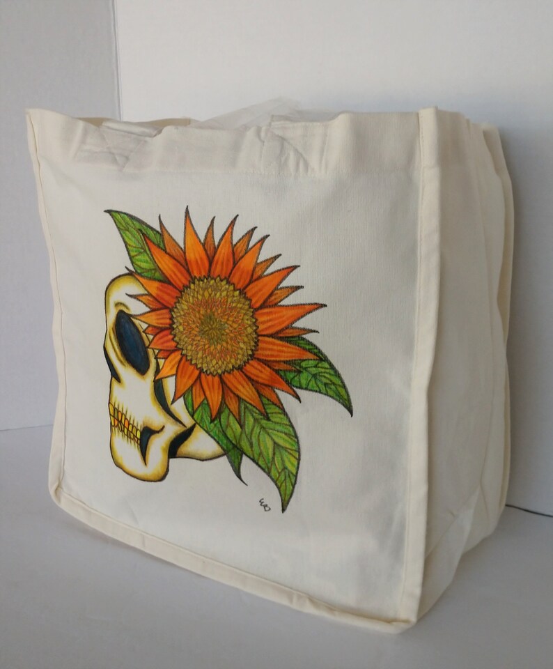 May include: A white canvas tote bag with a hand-painted design of a skull with a sunflower growing out of the top of its head. The sunflower is orange and yellow with green leaves.