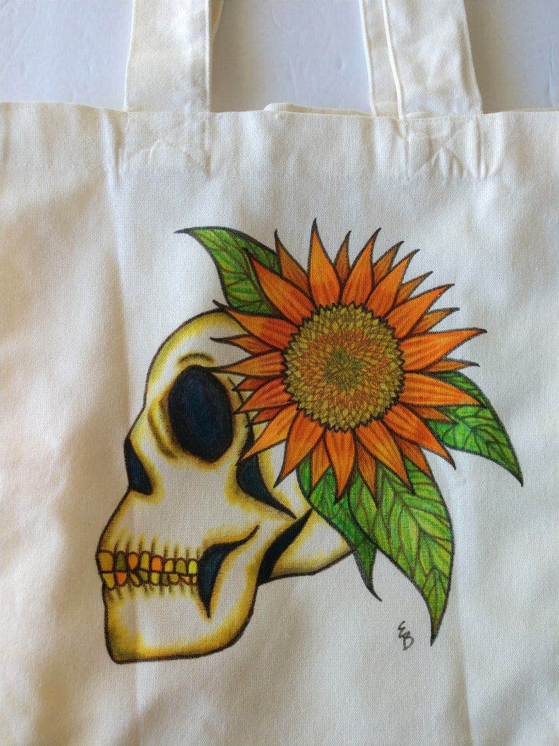 May include: A white tote bag with a hand-painted design of a skull wearing a sunflower crown. The skull is yellow and white with black eyes and a gold tooth. The sunflower is orange and yellow with green leaves. The bag is signed "WD".