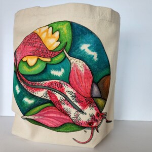 May include: A white canvas tote bag with a colorful hand-painted design of a pink and white fish with green and blue accents. The fish is swimming in a circle with a yellow flower.