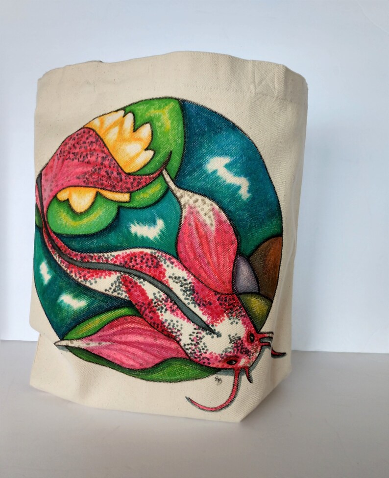 May include: A white canvas tote bag with a colorful hand-painted design of a pink and white fish with green and blue accents. The fish is swimming in a circle with a yellow flower.