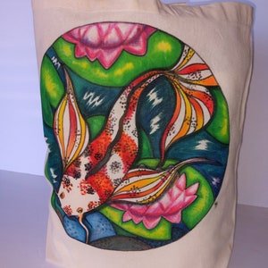 May include: A white canvas tote bag with a hand-painted design of a colorful koi fish swimming among pink water lilies and green lily pads. The design is in a circle on the front of the bag.