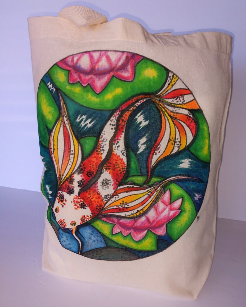 May include: A white canvas tote bag with a hand-painted design of a colorful koi fish swimming among pink water lilies and green lily pads. The design is in a circle on the front of the bag.