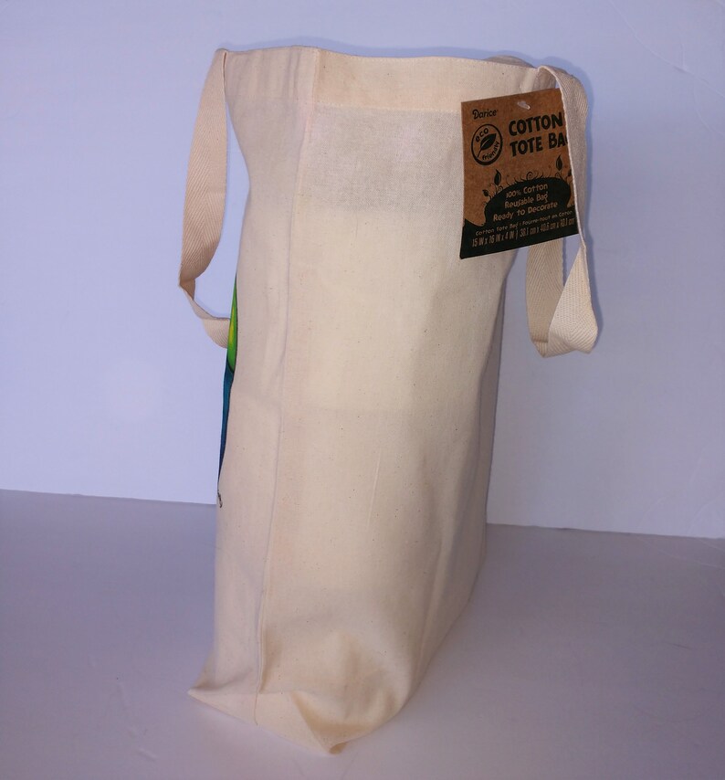 May include: A natural cotton tote bag with a brown paper tag that reads "Cotton Tote Bag 100% Cotton Reusable Bag Ready To Decorate".