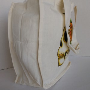 May include: A white canvas tote bag with a skull design featuring a sunflower on the side. The skull is yellow with black eyes and a gold tooth.