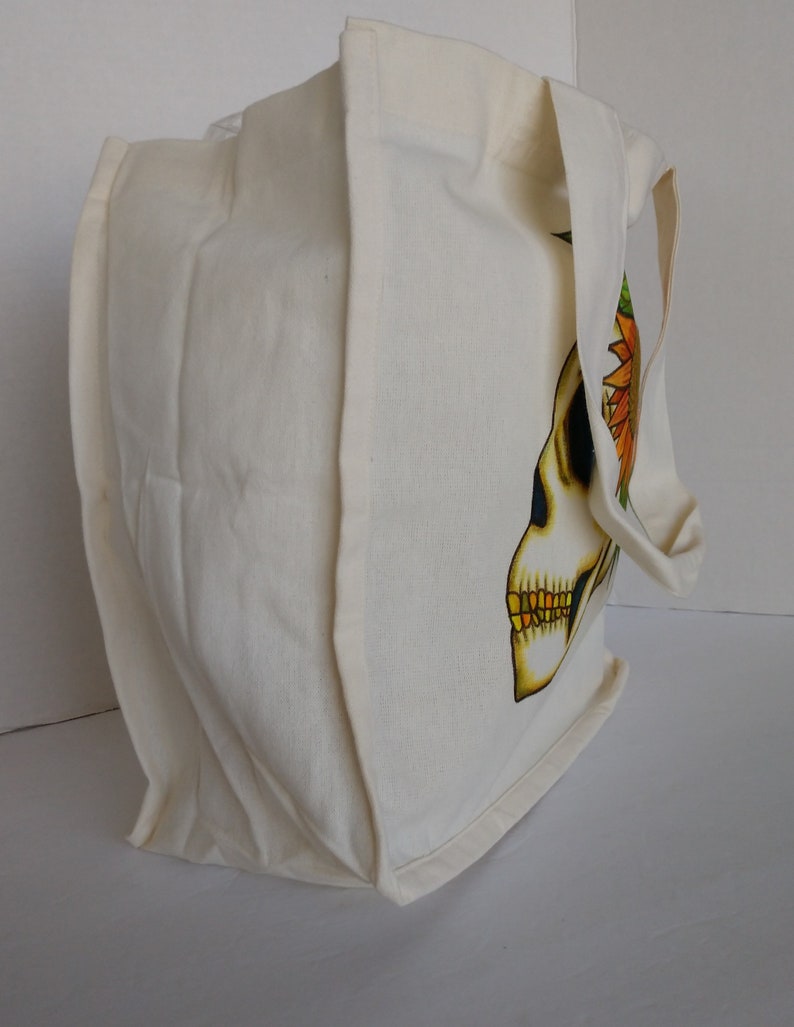 May include: A white canvas tote bag with a skull design featuring a sunflower on the side. The skull is yellow with black eyes and a gold tooth.