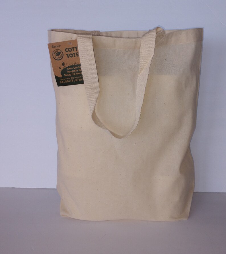 May include: A natural-colored cotton tote bag with a brown label that reads "Eco Cotton Tote Bag, 100% Cotton, Reusable Bag, Ready to Decorate, Carry Your Style, 10 oz, 14x16", and a green leaf logo.