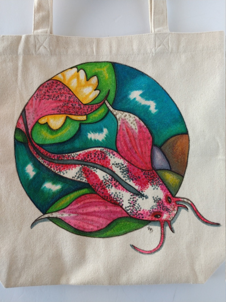 May include: A white canvas tote bag with a hand-painted design of a pink and white fish with black spots. The fish is surrounded by green, yellow, and blue shapes.