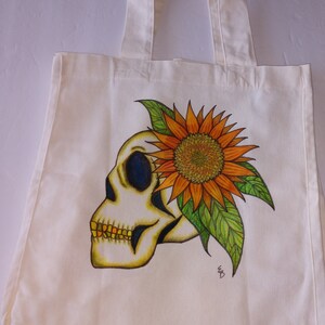May include: A white canvas tote bag with a hand-painted design of a skull with a sunflower growing out of the top of its head. The sunflower is orange and yellow with green leaves. The skull is white with black eyes and a yellow jaw.