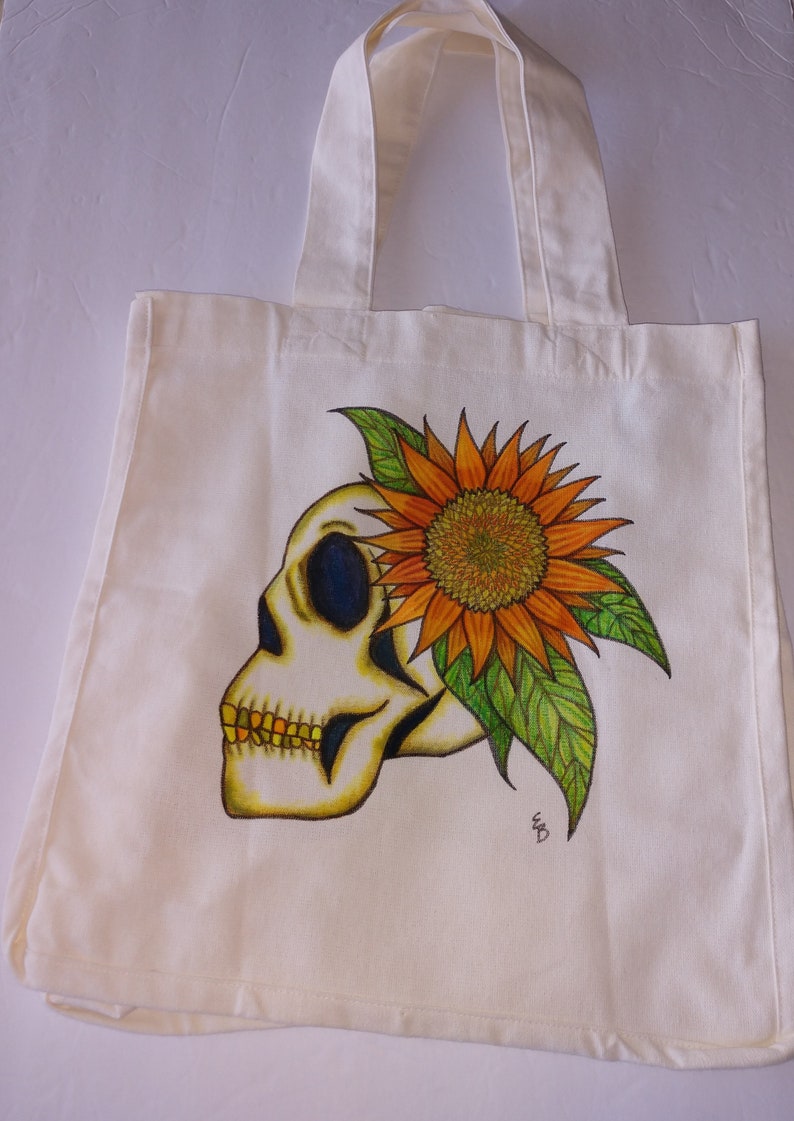 May include: A white canvas tote bag with a hand-painted design of a skull with a sunflower growing out of the top of its head. The sunflower is orange and yellow with green leaves. The skull is white with black eyes and a yellow jaw.