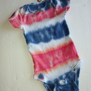 May include: A tie-dye baby onesie in shades of pink, white, and blue. The short-sleeved garment has a round neckline and snap closures at the bottom. The Gerber brand label is visible.