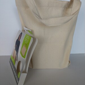 May include: A natural canvas tote bag with a large opening and two long straps. The bag is beige and has a slightly textured surface. The bag is sitting on a white surface. The bag is packaged with a cardboard insert that features a woman holding a similar bag and the text "Reusable & Sustainable Tote Bag. 100% Natural Cotton. Made in India. 100% Biodegradable. 100% Recyclable. 100% Eco-Friendly."