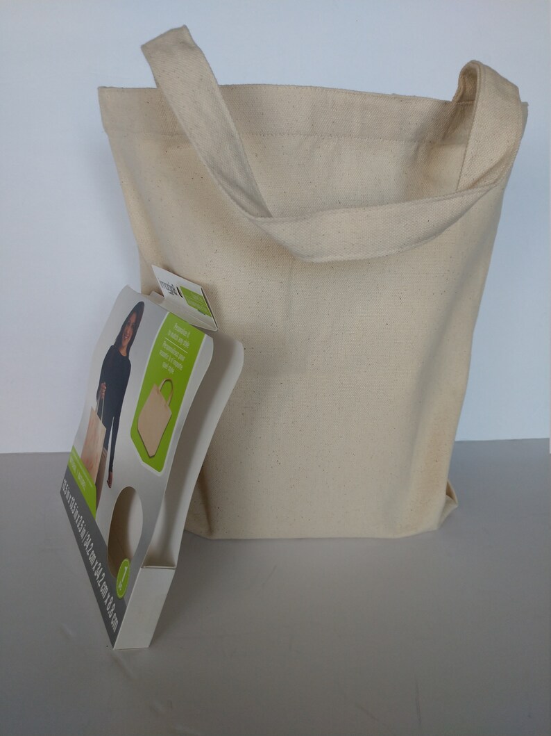 May include: A natural canvas tote bag with a large opening and two long straps. The bag is beige and has a slightly textured surface. The bag is sitting on a white surface. The bag is packaged with a cardboard insert that features a woman holding a similar bag and the text "Reusable & Sustainable Tote Bag. 100% Natural Cotton. Made in India. 100% Biodegradable. 100% Recyclable. 100% Eco-Friendly."