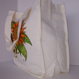 May include: A white canvas tote bag with a sunflower and skull design. The bag has two long straps and a large main compartment.