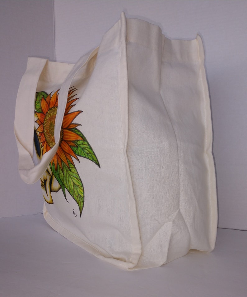 May include: A white canvas tote bag with a sunflower and skull design. The bag has two long straps and a large main compartment.