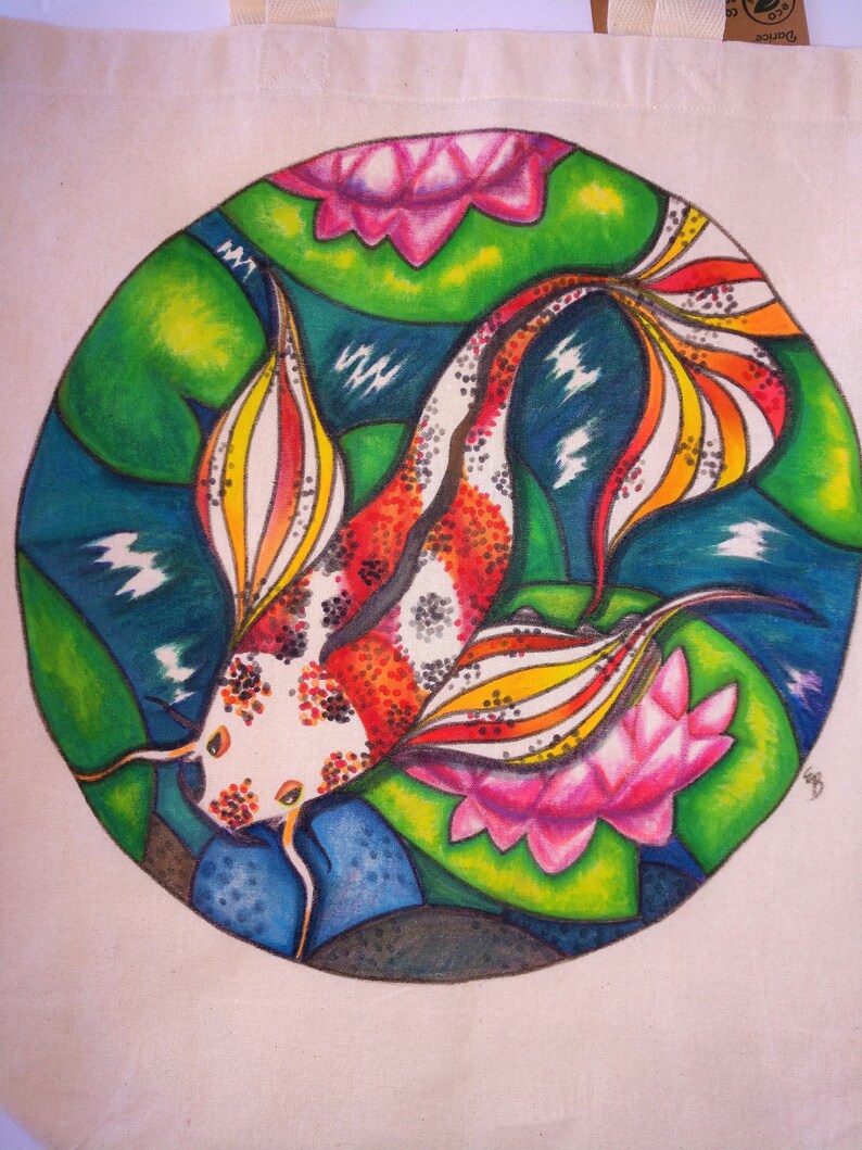 May include: A white tote bag with a colorful hand-painted design of a koi fish swimming in a pond with lily pads. The fish is red, orange, yellow, and black with white spots. The lily pads are pink and green.