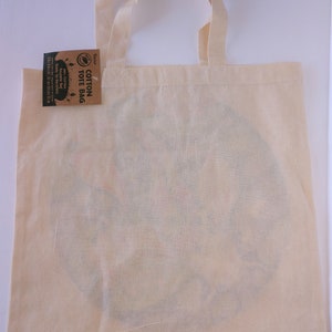 May include: A natural cotton tote bag with a brown tag that reads "Cotton Tote Bag" and has a leaf design. The bag is blank and ready to be decorated.