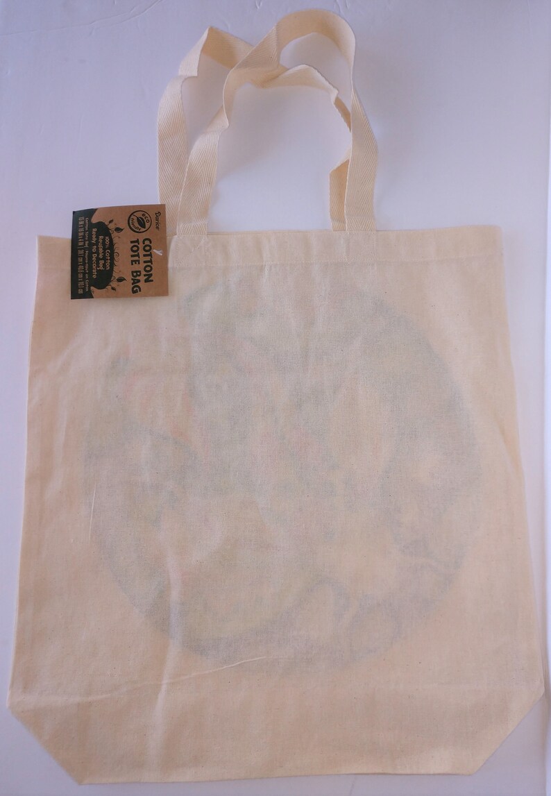 May include: A natural cotton tote bag with a brown tag that reads "Cotton Tote Bag" and has a leaf design. The bag is blank and ready to be decorated.