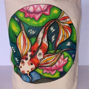 May include: A white canvas tote bag with a colorful hand-painted design of a koi fish swimming among pink and green water lilies. The fish has red, orange, yellow, black, and white scales.