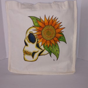 May include: A white canvas tote bag with a hand-painted design of a skull with a sunflower on top. The skull is white with black eyes and a yellow mouth. The sunflower is orange and yellow with green leaves.