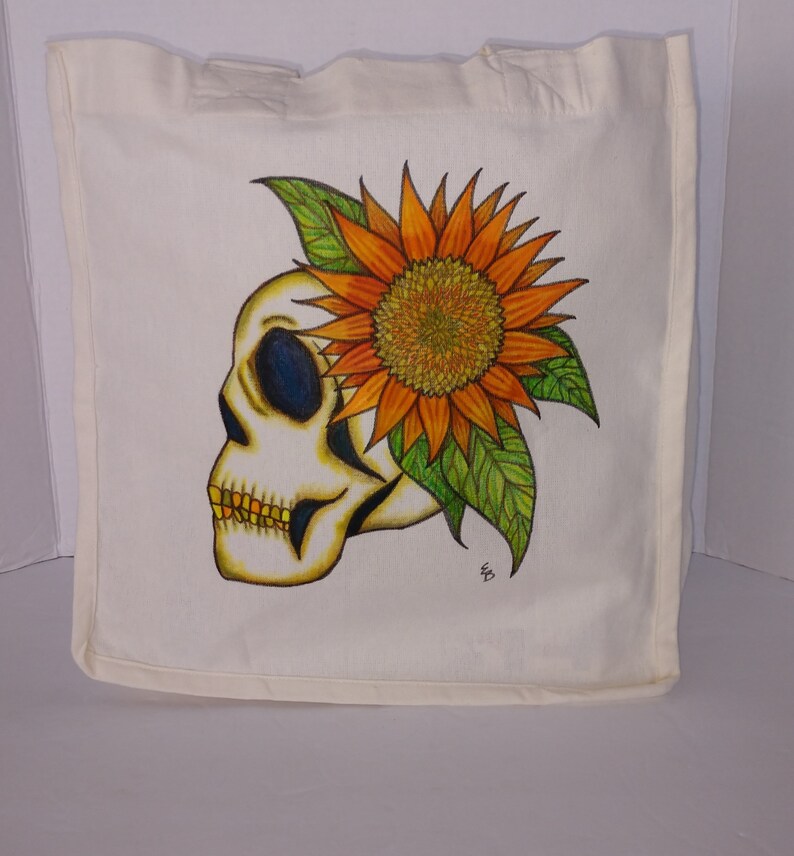 May include: A white canvas tote bag with a hand-painted design of a skull with a sunflower on top. The skull is white with black eyes and a yellow mouth. The sunflower is orange and yellow with green leaves.